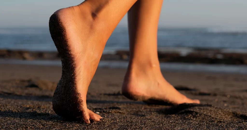 Pés descalços caminhando na areia da praia ao pôr do sol, representando bem-estar e saúde dos pés.